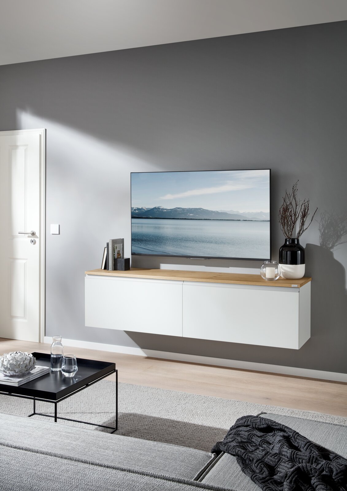 Modernes, schwebendes TV-Lowboard in Weiß mit Holzplatte unter einem Flachbildfernseher, dekoriert mit Büchern, Kerze und Vase, in einem hellen Wohnzimmer – Frontansicht in leichter Schrägperspektive von links.
