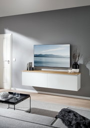 Modernes, schwebendes TV-Lowboard in Weiß mit Holzplatte unter einem Flachbildfernseher, dekoriert mit Büchern, Kerze und Vase, in einem hellen Wohnzimmer – Frontansicht in leichter Schrägperspektive von links.