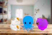 Vorderansicht von drei leuchtenden Nachtlichtern in Tierform: eine weiße Katze mit orangefarbenem Fleck, ein blaues Oktopus und ein pinker Bär, auf einem Holztisch in einem Kinderzimmer.