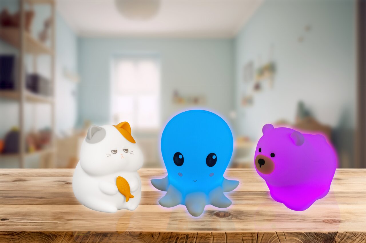 Vorderansicht von drei leuchtenden Nachtlichtern in Tierform: eine weiße Katze mit orangefarbenem Fleck, ein blaues Oktopus und ein pinker Bär, auf einem Holztisch in einem Kinderzimmer.