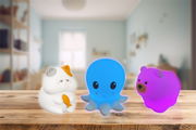 Vorderansicht von drei leuchtenden Nachtlichtern in Tierform: eine weiße Katze mit orangefarbenem Fleck, ein blaues Oktopus und ein pinker Bär, auf einem Holztisch in einem Kinderzimmer.