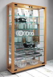 Moderne Glasvitrine mit hellem Holzrahmen, Glasböden und Glastüren, gefüllt mit Deko und Büchern; schräg von vorne links aufgenommen.