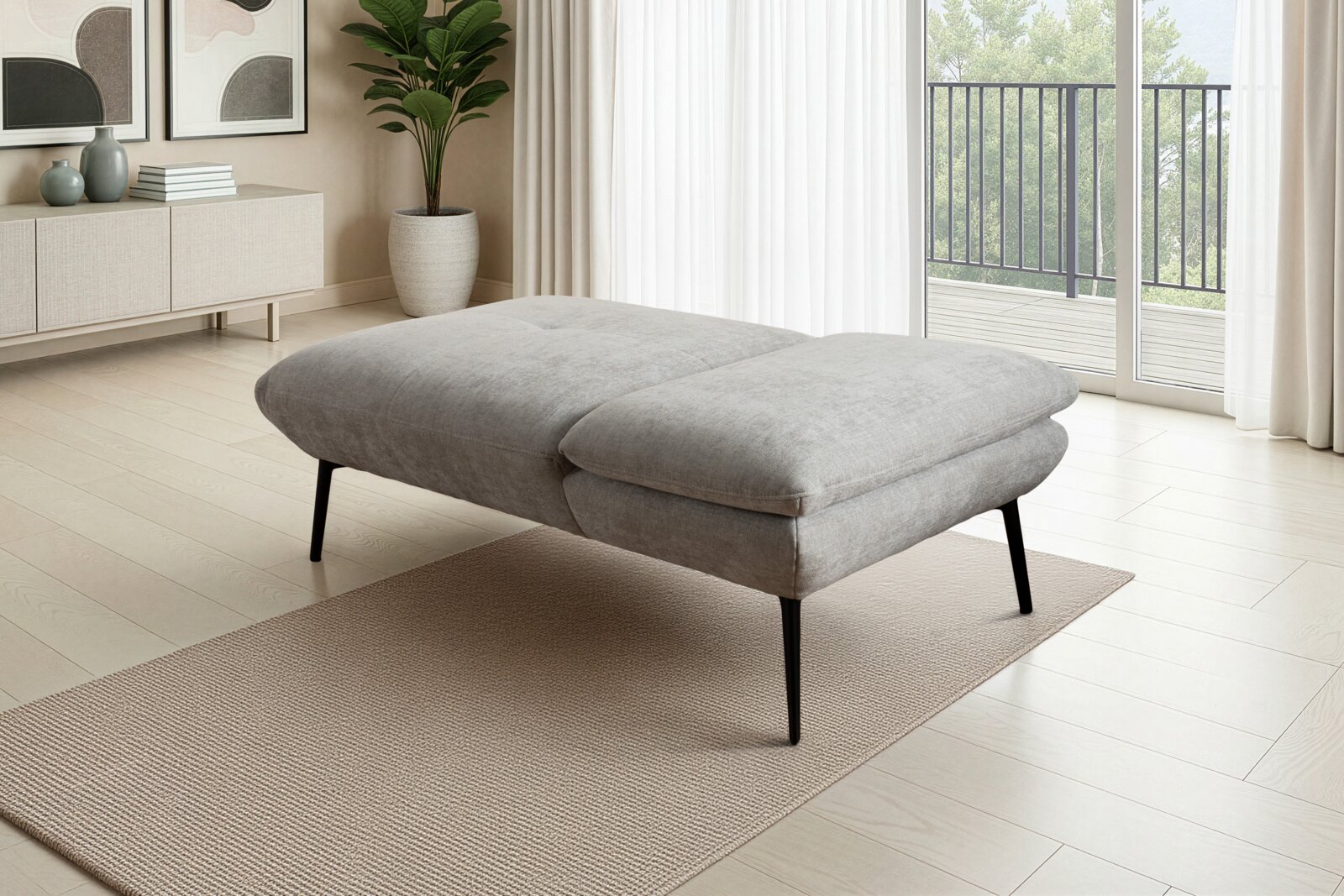 Grijze, gestoffeerde poef/daybed met zwarte metalen poten op een beige tapijt in een lichte woonkamer, gefotografeerd vanuit een zijaanzicht (licht van voren).