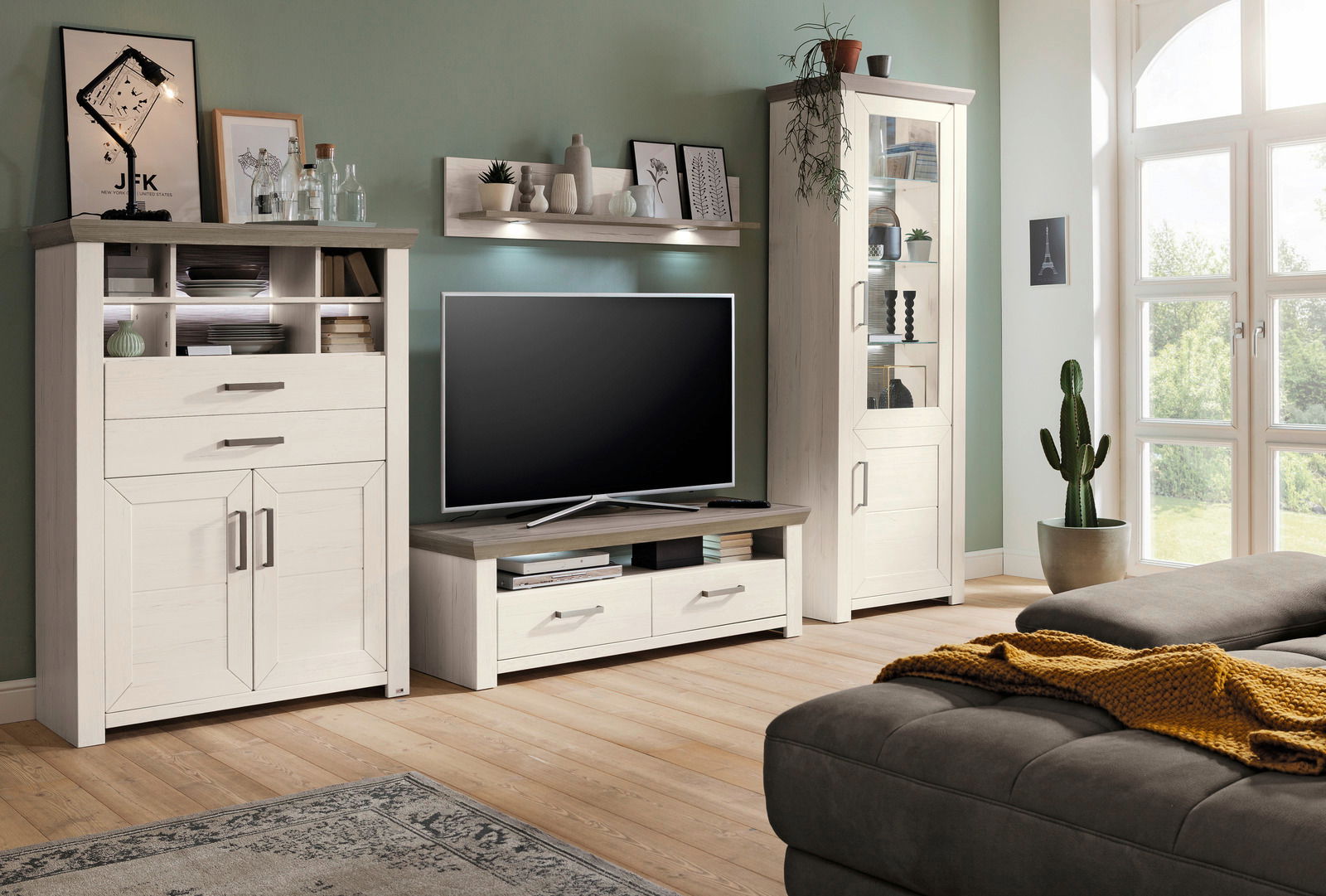 Set one by Musterring Vitrine SET ONE YORK Wohnzimmermöbel-Set in heller Holzoptik mit Highboard, Lowboard und Vitrine, dekoriert mit Pflanzen und Bildern, seitliche Perspektive.