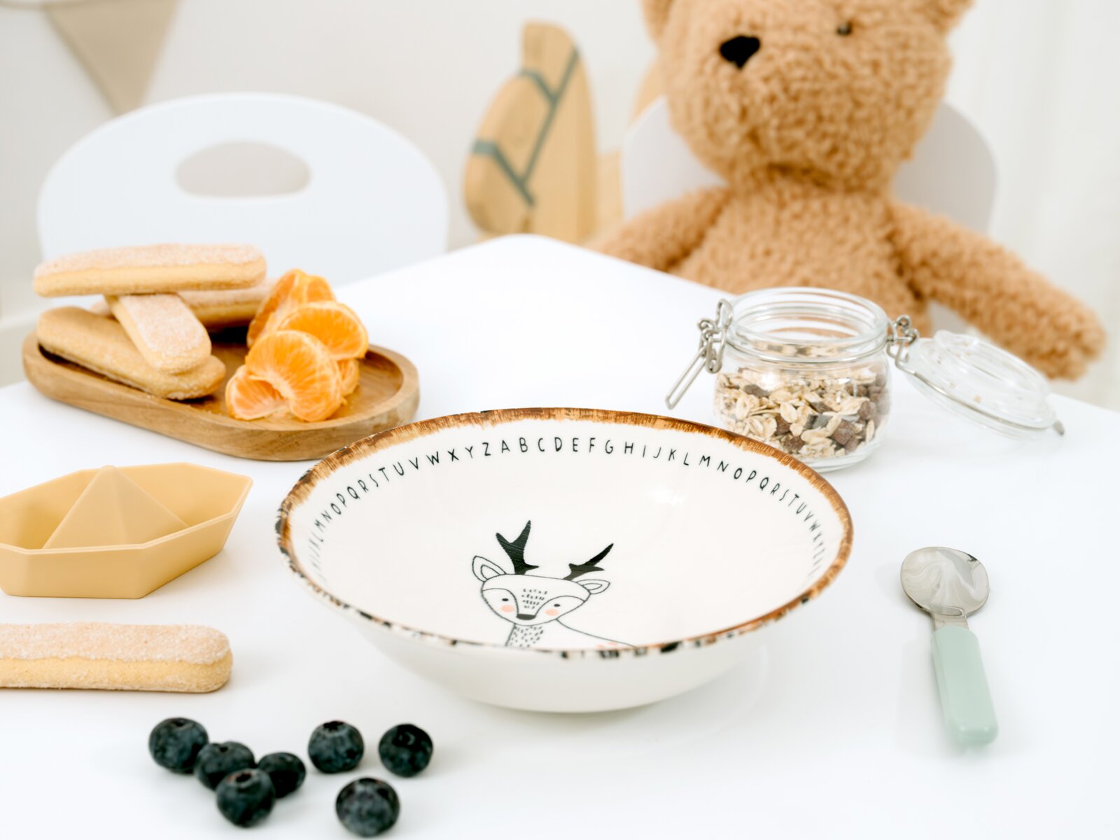 Weiße Kinderschale mit niedlichem Reh- und Alphabetmotiv sowie braunem Rand, aus schräger Vorderansicht von oben in einer liebevoll arrangierten Frühstücksszene gezeigt.