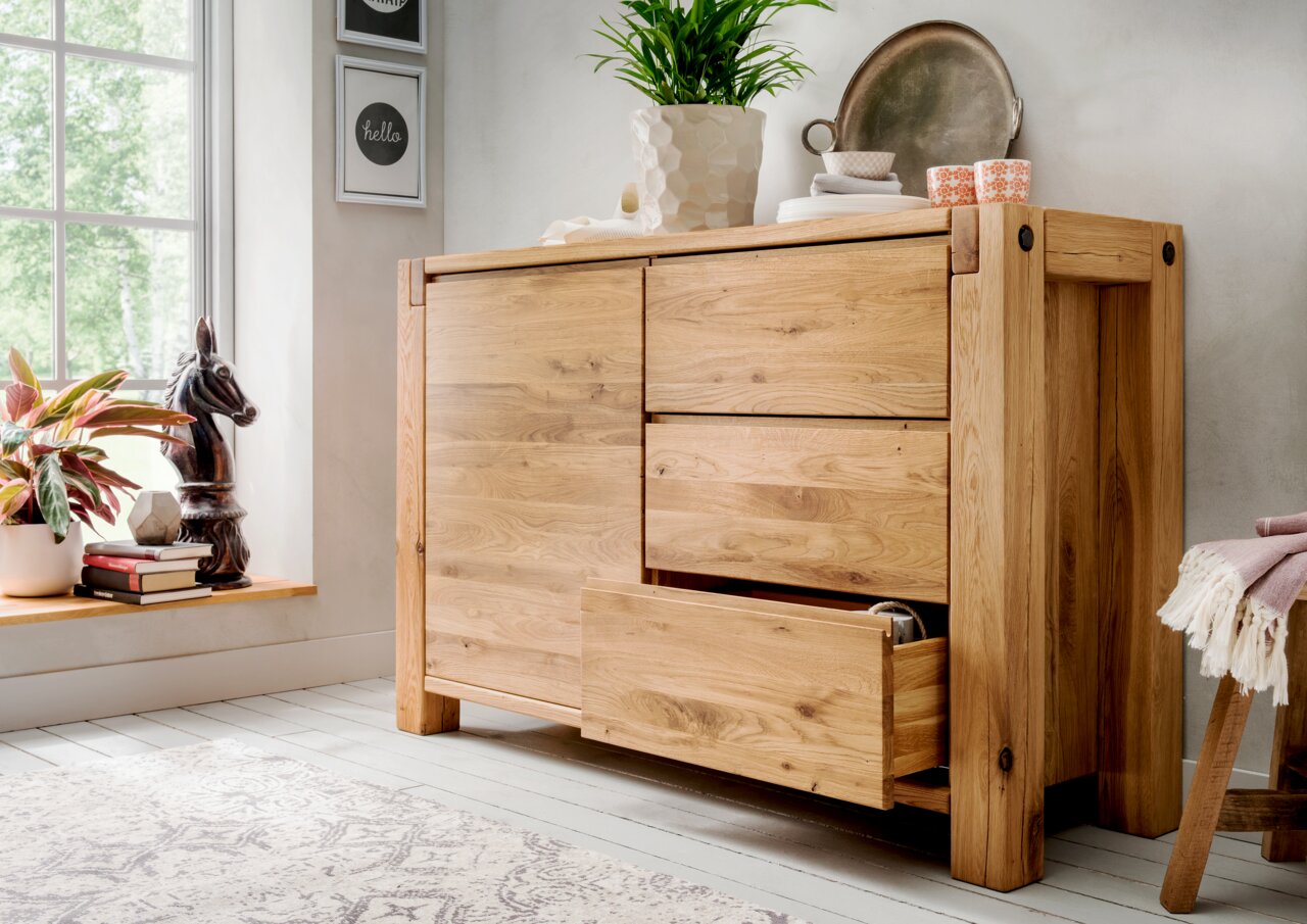 Holzkommode mit zwei Türen und zwei Schubladen, teilweise geöffnet, in einem hellen Raum mit Pflanzen und Dekorationen, seitliche Perspektive