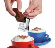 Mini-Vierkantreibe in der Hand, die Schokolade über einer Tasse mit Milchschaum reibt, seitliche Perspektive.
