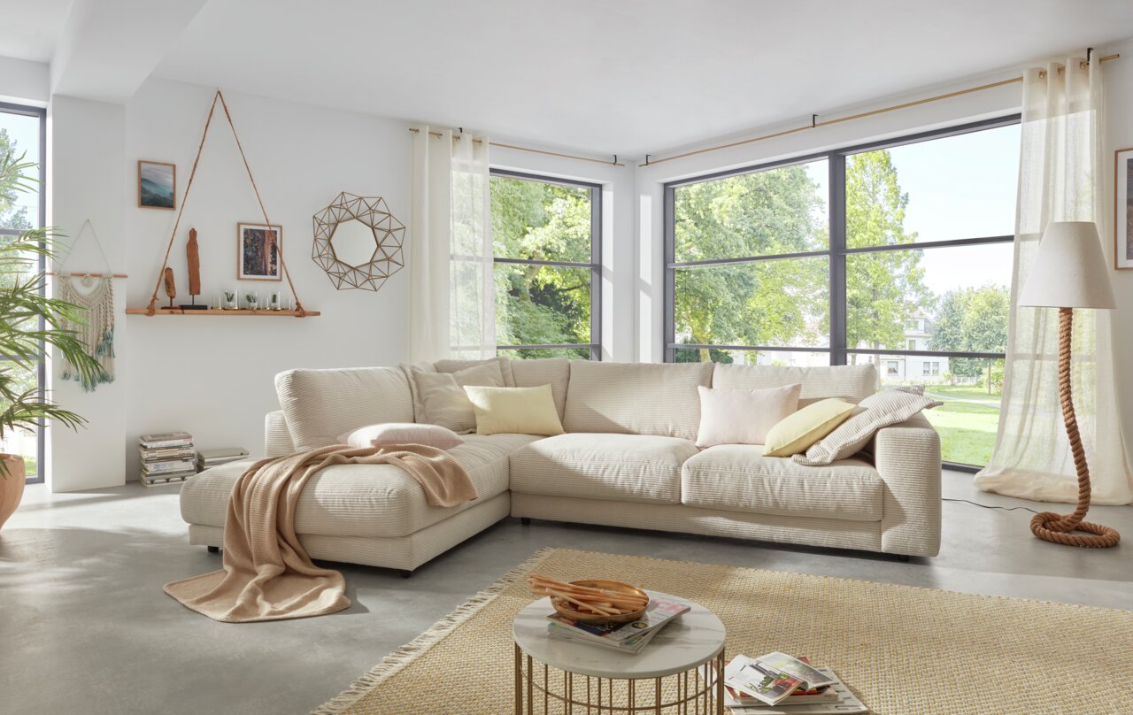 Ecksofa Seventies Gemütliche Rundecke in einem modernen Wohnzimmer mit großen Fenstern, die viel Tageslicht hereinlassen. Die Perspektive zeigt die Rundecke von vorne, umgeben von dekorativen Kissen und einer Decke.