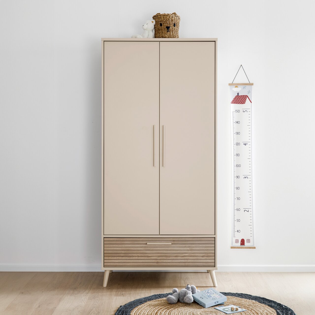 Moderner Kleiderschrank mit zwei Türen und einer Schublade in hellem Holzdesign, frontal fotografiert. Dekoriert mit einem Korb und einem Stofftier auf der Oberseite.