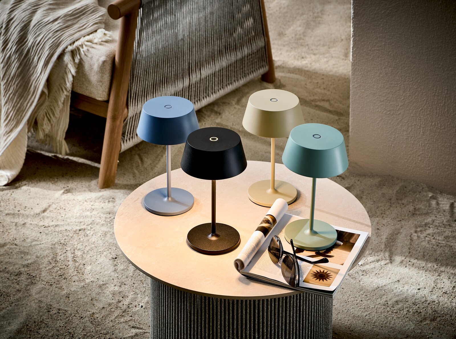 Vier moderne kabellose LED-Tischlampen in Blau, Schwarz, Creme und Mint auf rundem Couchtisch neben Magazin und Sonnenbrille, schräg von oben fotografiert.