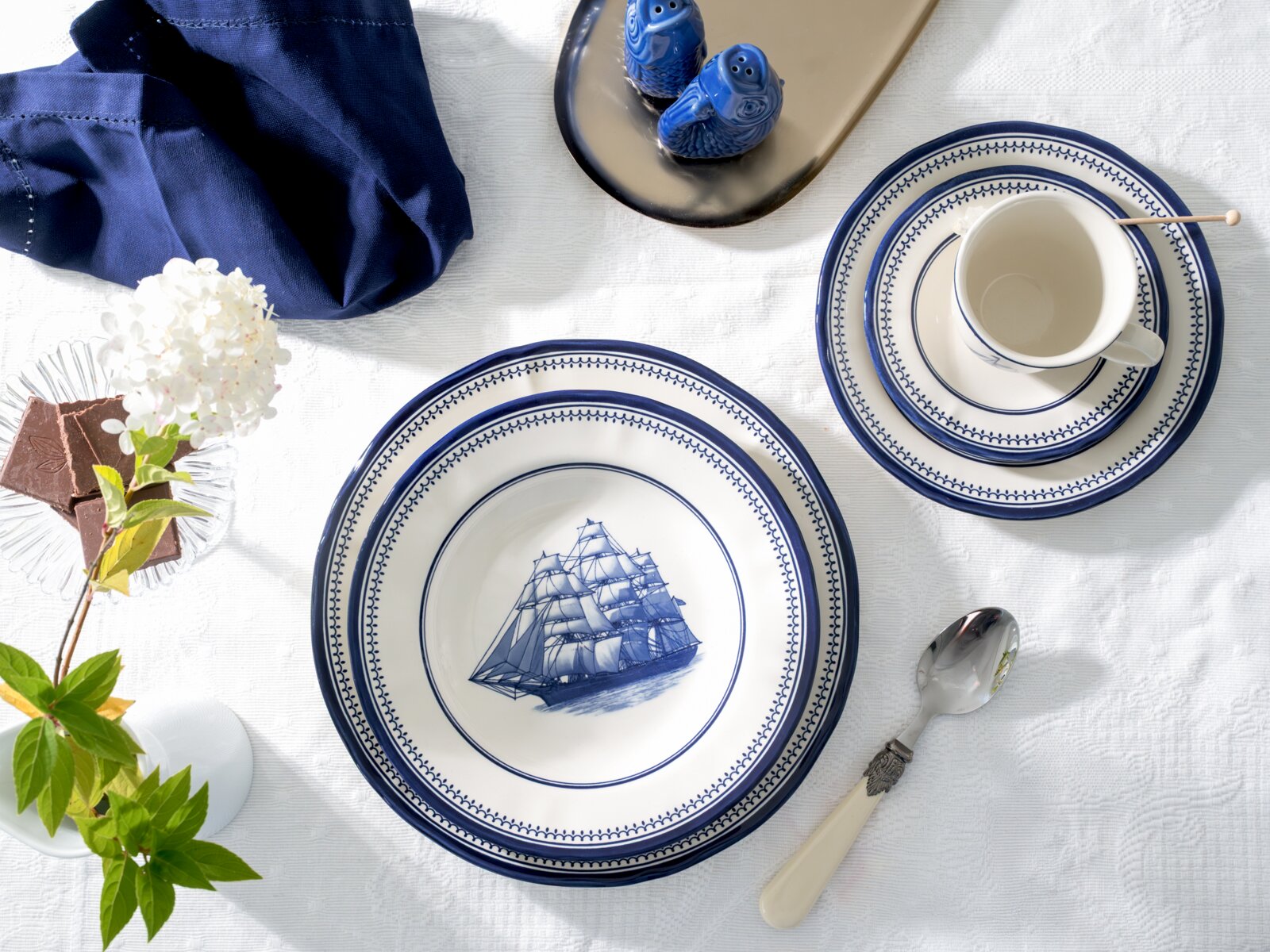 Maritimes Porzellan-Geschirrset in Weiß mit kobaltblauem Randdekor und Segelschiff-Motiv, arrangiert auf weißer Tischdecke mit Tasse, Tellern und Löffel, Draufsicht von oben