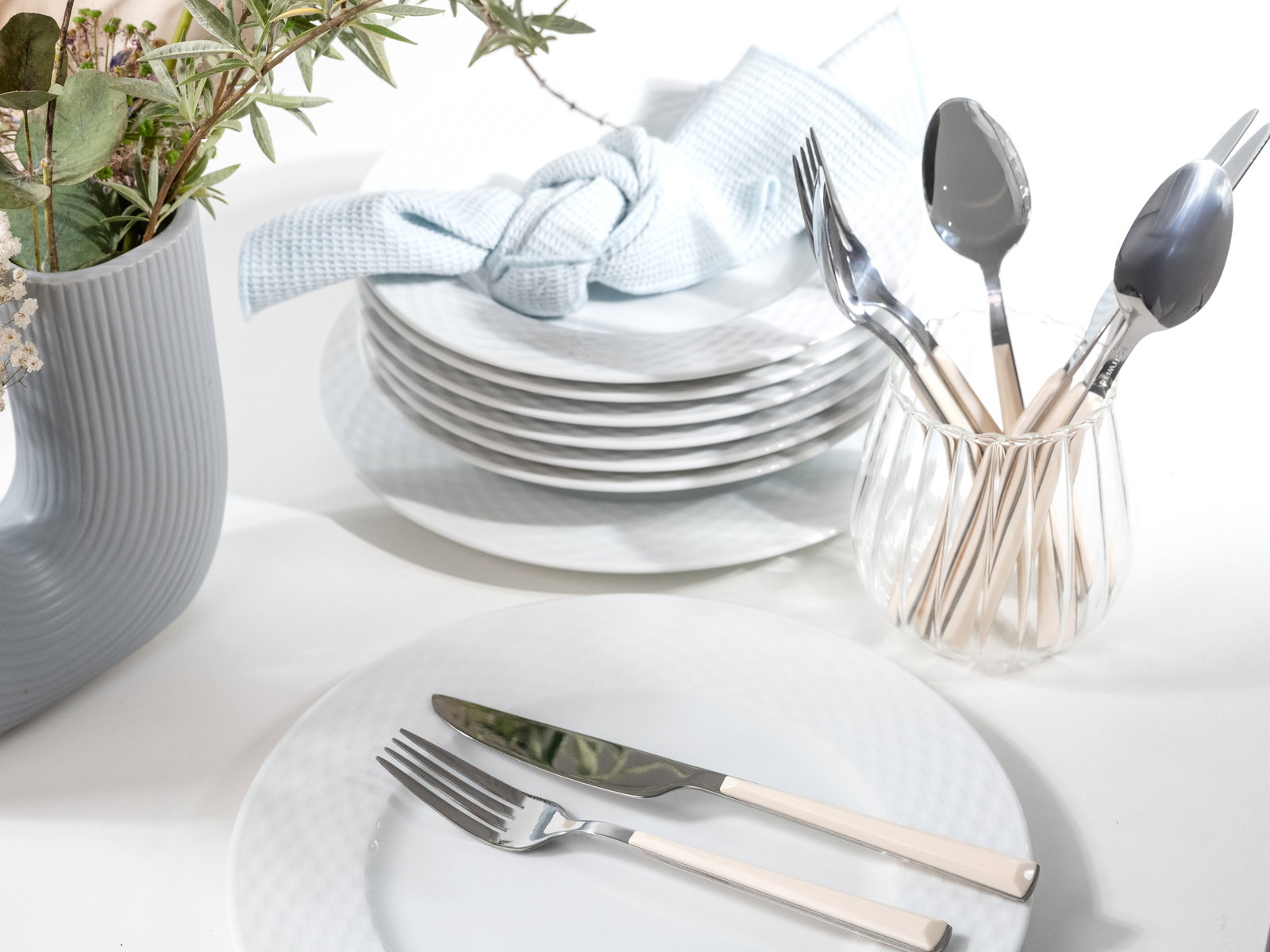 Elegantes Tafelservice mit gestapelten weißen Tellern, Besteck in einem Glas und einer blauen Serviette, aus der Vogelperspektive fotografiert.