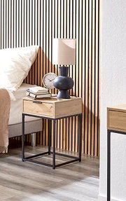 Moderner Nachttisch aus hellem Holz mit Schublade und schwarzem Metallgestell neben einem Bett, darauf Tischlampe, Wecker und Bücher, seitliche Perspektive