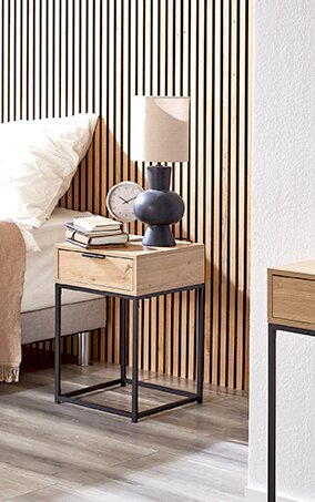 Moderner Nachttisch aus hellem Holz mit Schublade und schwarzem Metallgestell neben einem Bett, darauf Tischlampe, Wecker und Bücher, seitliche Perspektive
