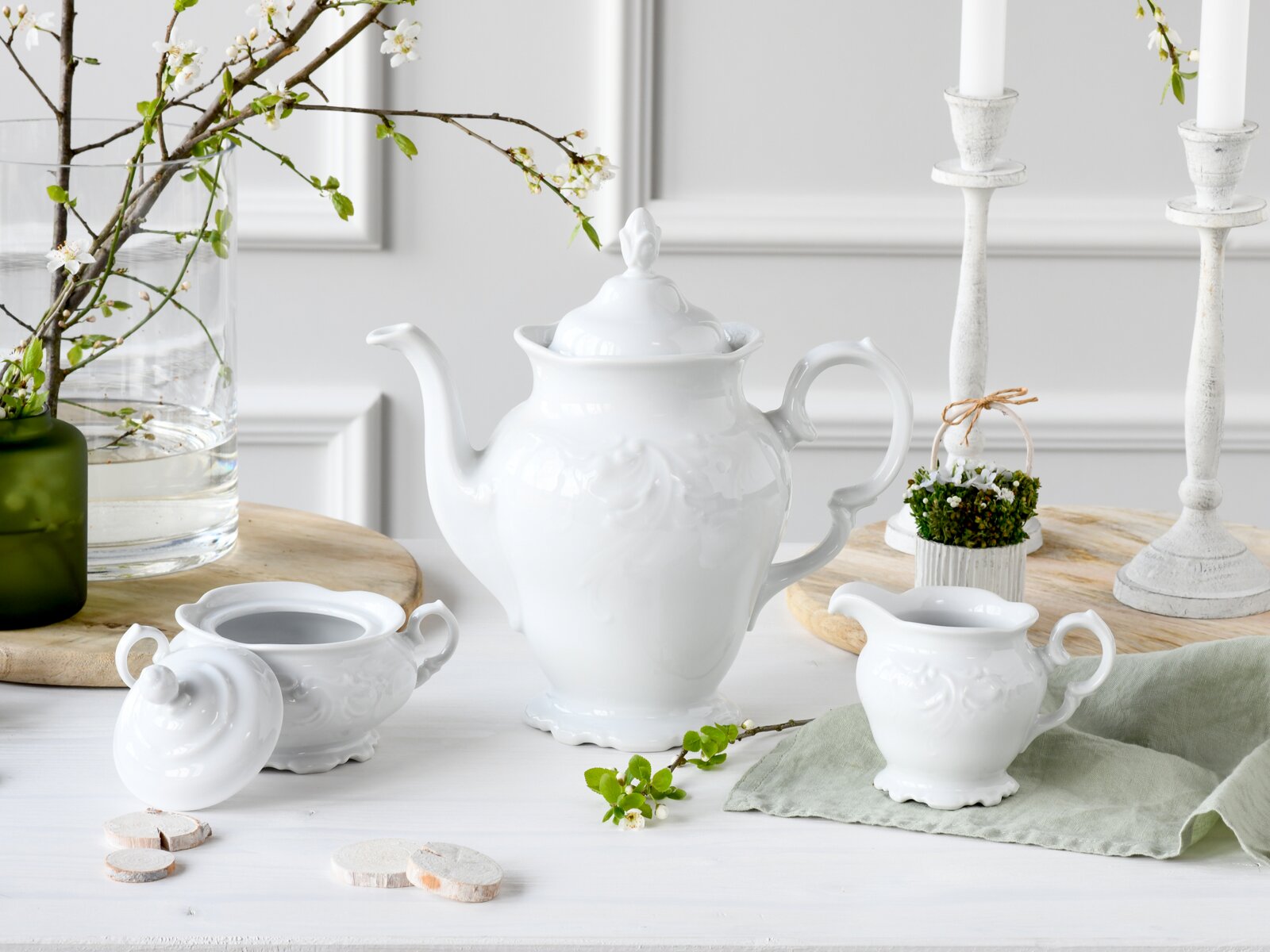 Frontalansicht eines eleganten weißen Porzellan-Teeservices mit großer Teekanne, Zuckerdose und Milchkännchen auf hellem Tisch, dekoriert mit Frühlingszweigen und Kerzenständern im Hintergrund.