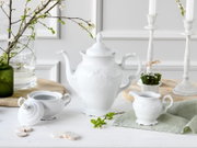 Frontalansicht eines eleganten weißen Porzellan-Teeservices mit großer Teekanne, Zuckerdose und Milchkännchen auf hellem Tisch, dekoriert mit Frühlingszweigen und Kerzenständern im Hintergrund.
