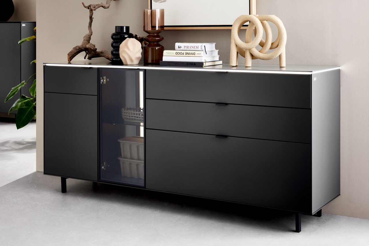 Modernes schwarzes Sideboard mit Schubladen und Glastür, dekoriert mit Büchern und Skulpturen, seitliche Perspektive