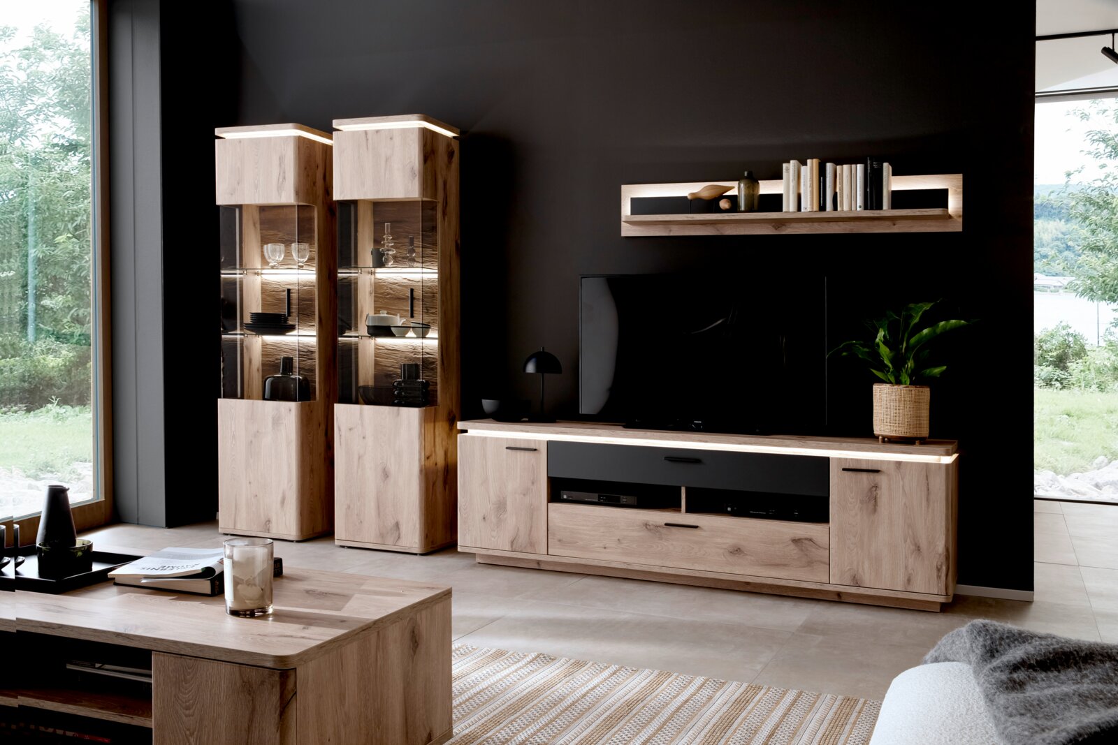 Zurbrüggen Wandcombinatie MODENA Moderne entertainmentset voor de woonkamer van licht hout, bestaande uit een tv-lowboard, twee verlichte glazen vitrinekasten en een wandplank voor een donkere muur, gefotografeerd van voren vanuit een licht zijdelings perspectief