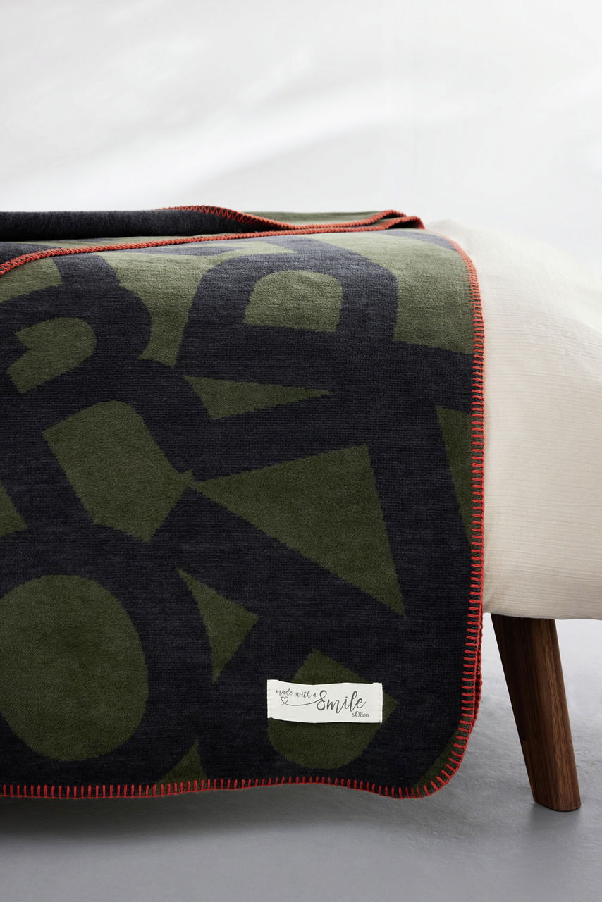 Jacquard Decke von s.Oliver in Olivgrün und Schwarz mit rotem Rand, auf einem Bett drapiert, seitliche Perspektive