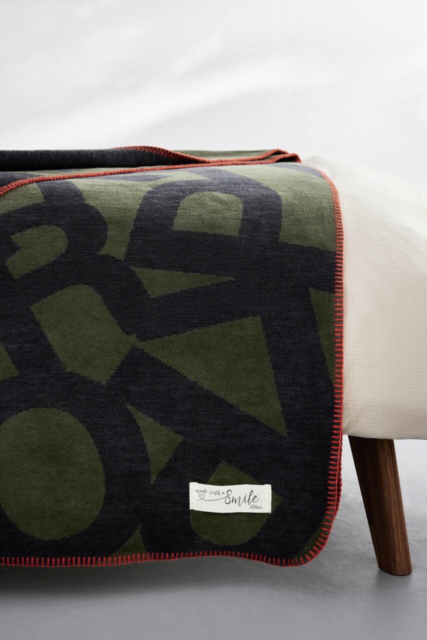 Jacquarddeken van s.Oliver in olijfgroen en zwart met een rode rand, gedrapeerd over een bed, zijaanzicht