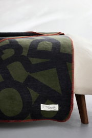 Jacquarddeken van s.Oliver in olijfgroen en zwart met een rode rand, gedrapeerd over een bed, zijaanzicht