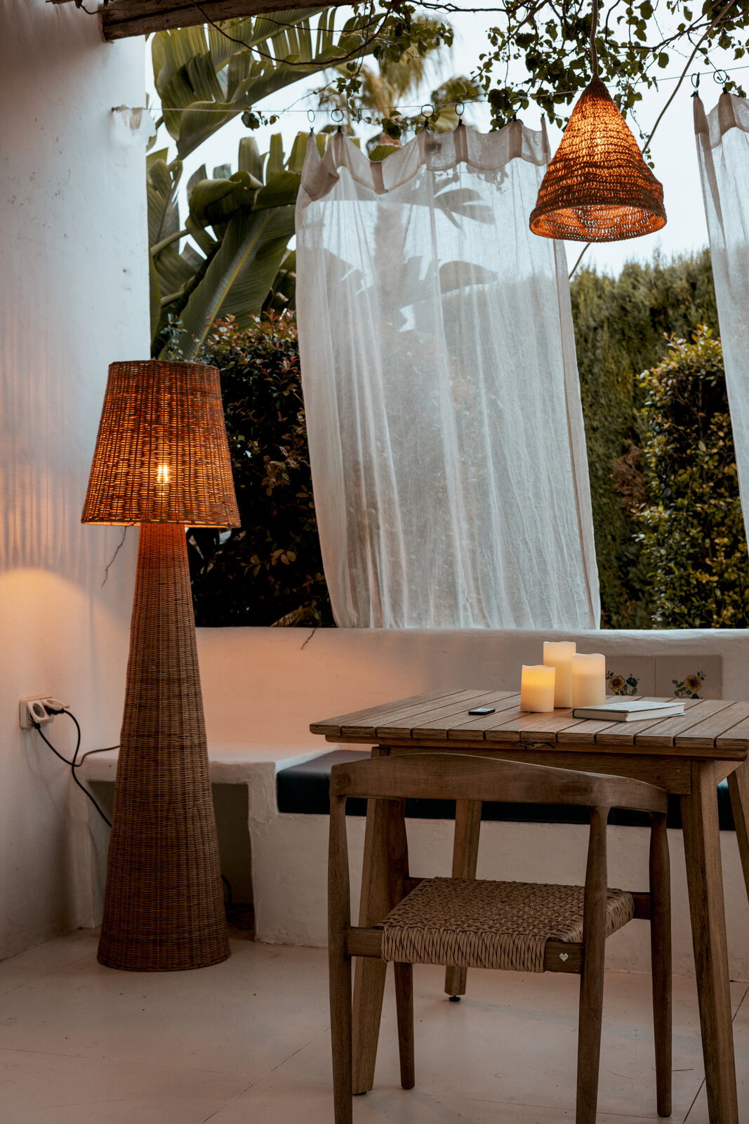 Gemütliche Outdoor-Terrasse mit geflochtener Rattan-Stehlampe und hängender Korblampe, Holztisch mit Kerzen und Stuhl, aufgenommen aus leicht seitlicher Perspektive auf Augenhöhe.