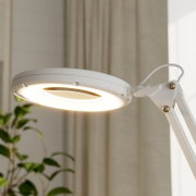 Weiße LED-Schreibtischlampe mit rundem Leuchtkopf und warmem Licht, Detailaufnahme aus seitlicher Untersicht in einem Wohnraum mit Vorhängen und Pflanze im Hintergrund