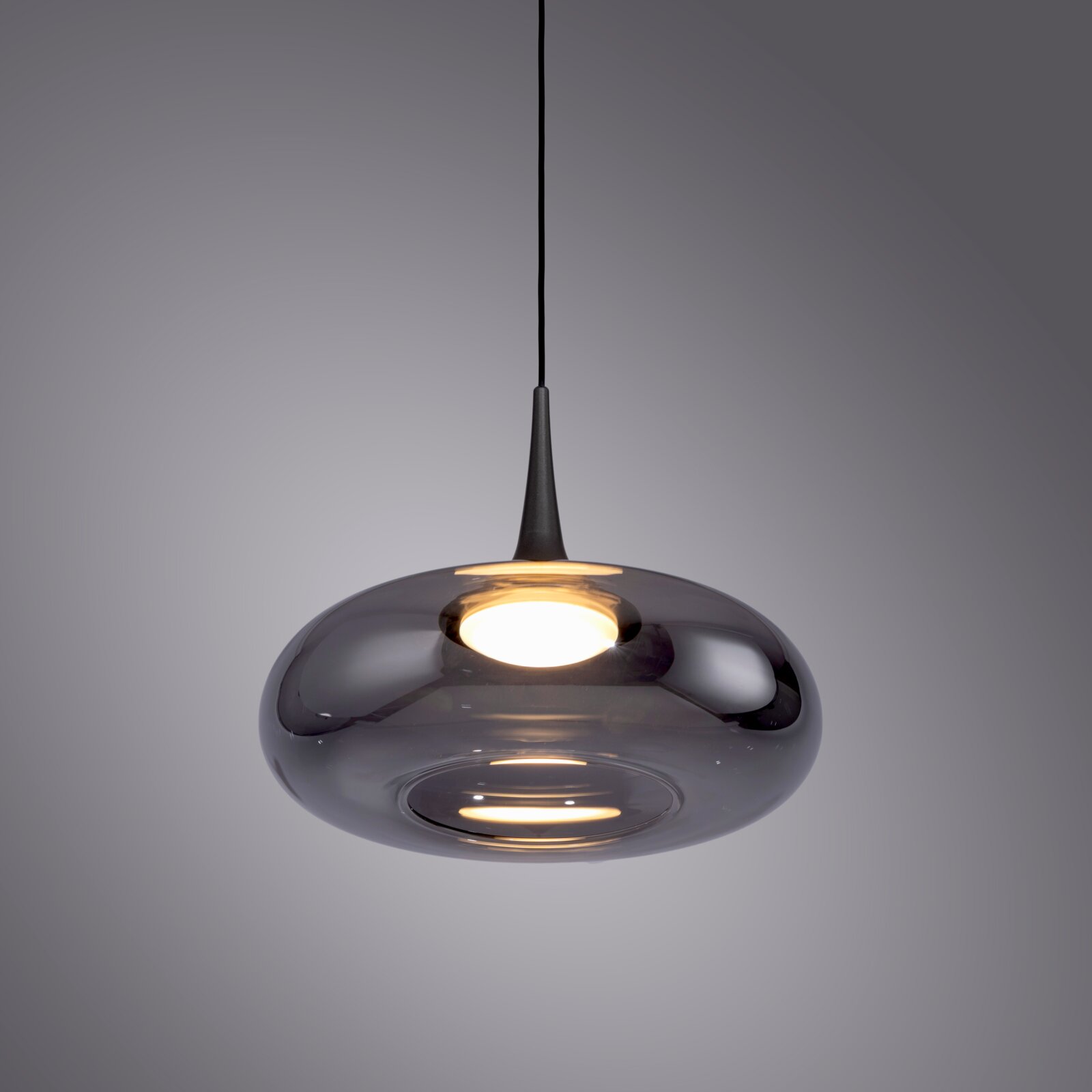Moderne Pendelleuchte mit rauchgrauem, glänzendem Glasschirm in flacher, runder Form und schwarzer Aufhängung, eingeschaltet vor grauem Hintergrund, Ansicht von schräg unten