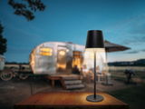 Moderne Akku-Tischleuchte in schwarzem Design auf einem Holztisch, mit einem unscharfen Wohnwagen im Hintergrund, aus frontaler Perspektive fotografiert.
