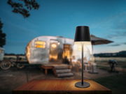 Moderne Akku-Tischleuchte in schwarzem Design auf einem Holztisch, mit einem unscharfen Wohnwagen im Hintergrund, aus frontaler Perspektive fotografiert.