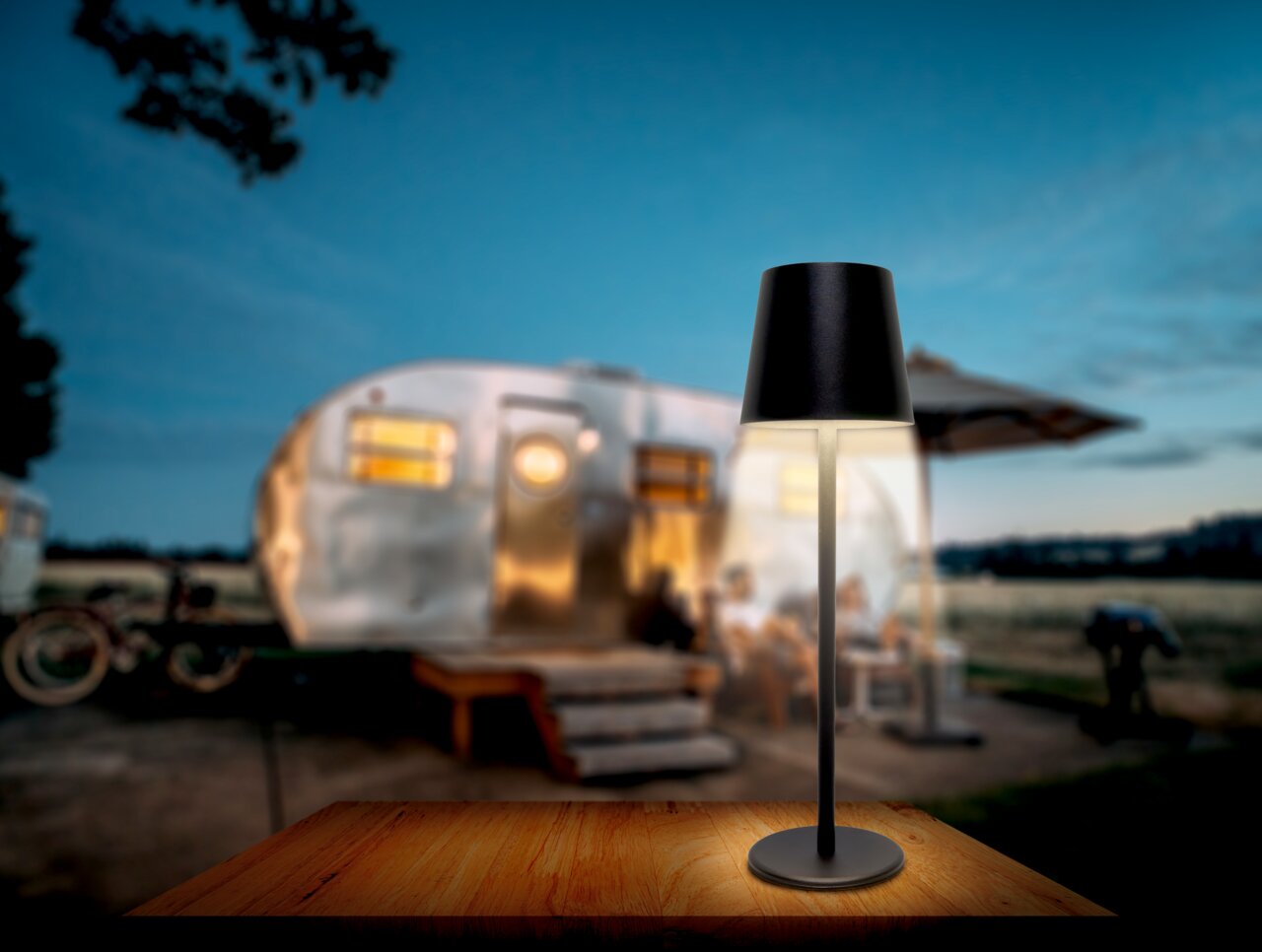Moderne Akku-Tischleuchte in schwarzem Design auf einem Holztisch, mit einem unscharfen Wohnwagen im Hintergrund, aus frontaler Perspektive fotografiert.
