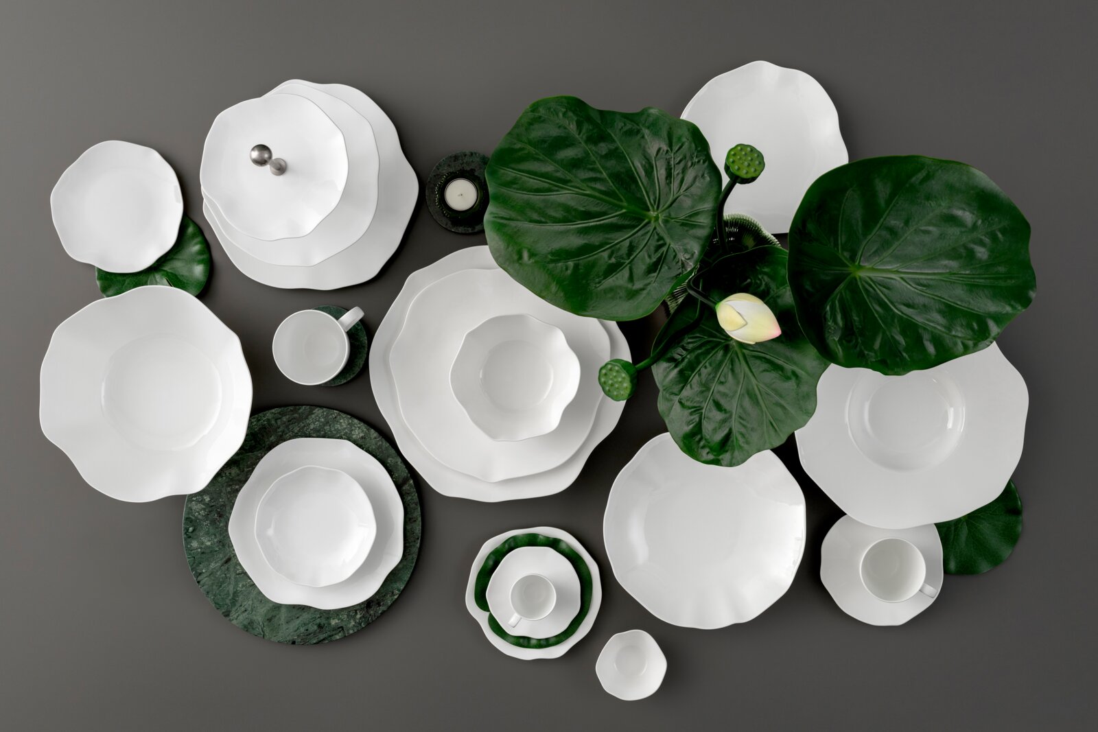 Draufsicht (Flatlay) auf ein elegantes weißes Porzellan-Geschirrset mit Tellern, Schalen und Tassen auf grauem Hintergrund, dekoriert mit großen grünen Blättern und einer hellen Blüte.