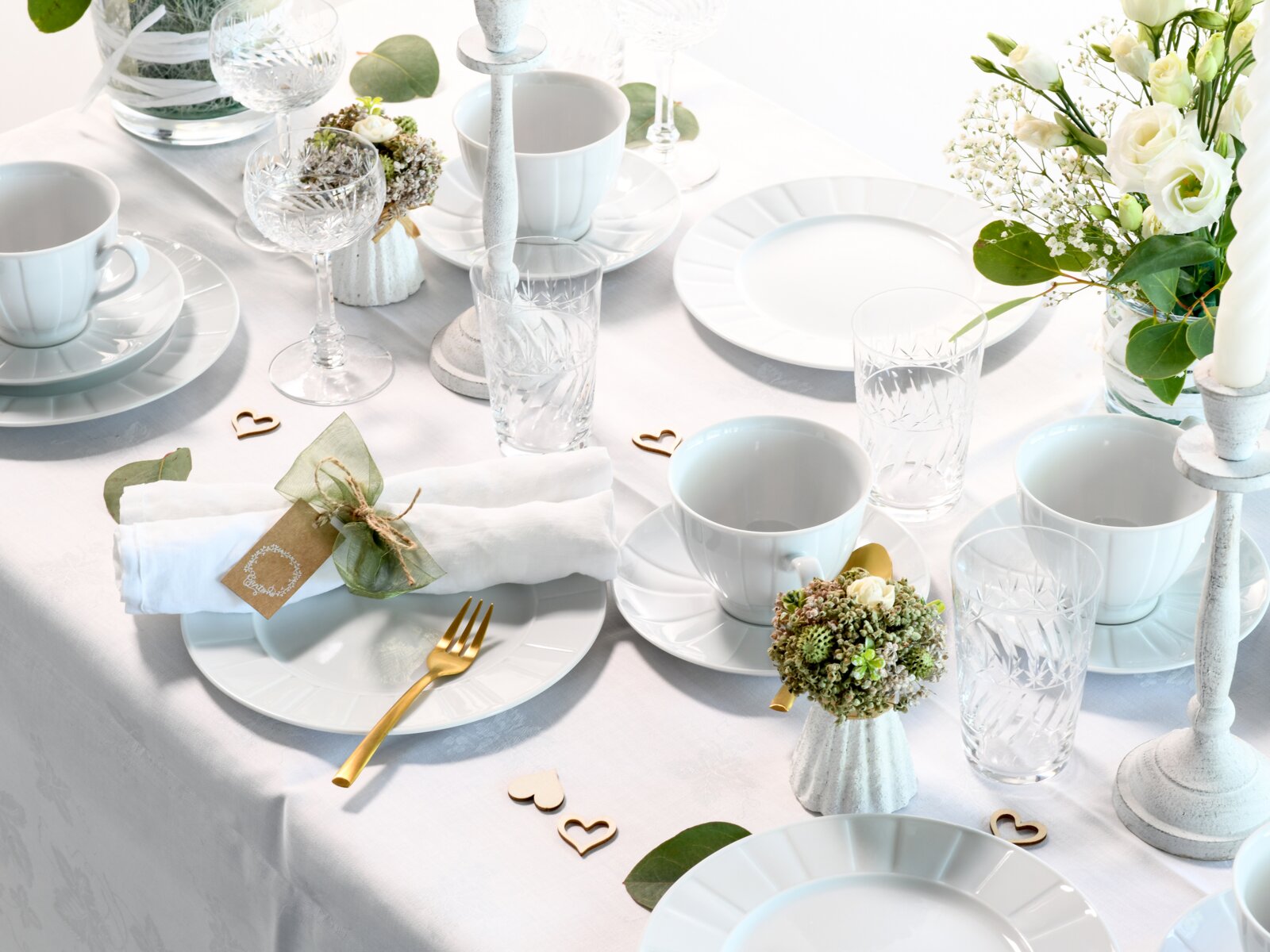 Festlich gedeckter Tisch mit weißem Porzellangeschirr, Kristallgläsern, goldfarbenem Besteck, Kerzenständern und weiß-grüner Blumendeko, aus schräger Vogelperspektive von oben fotografiert