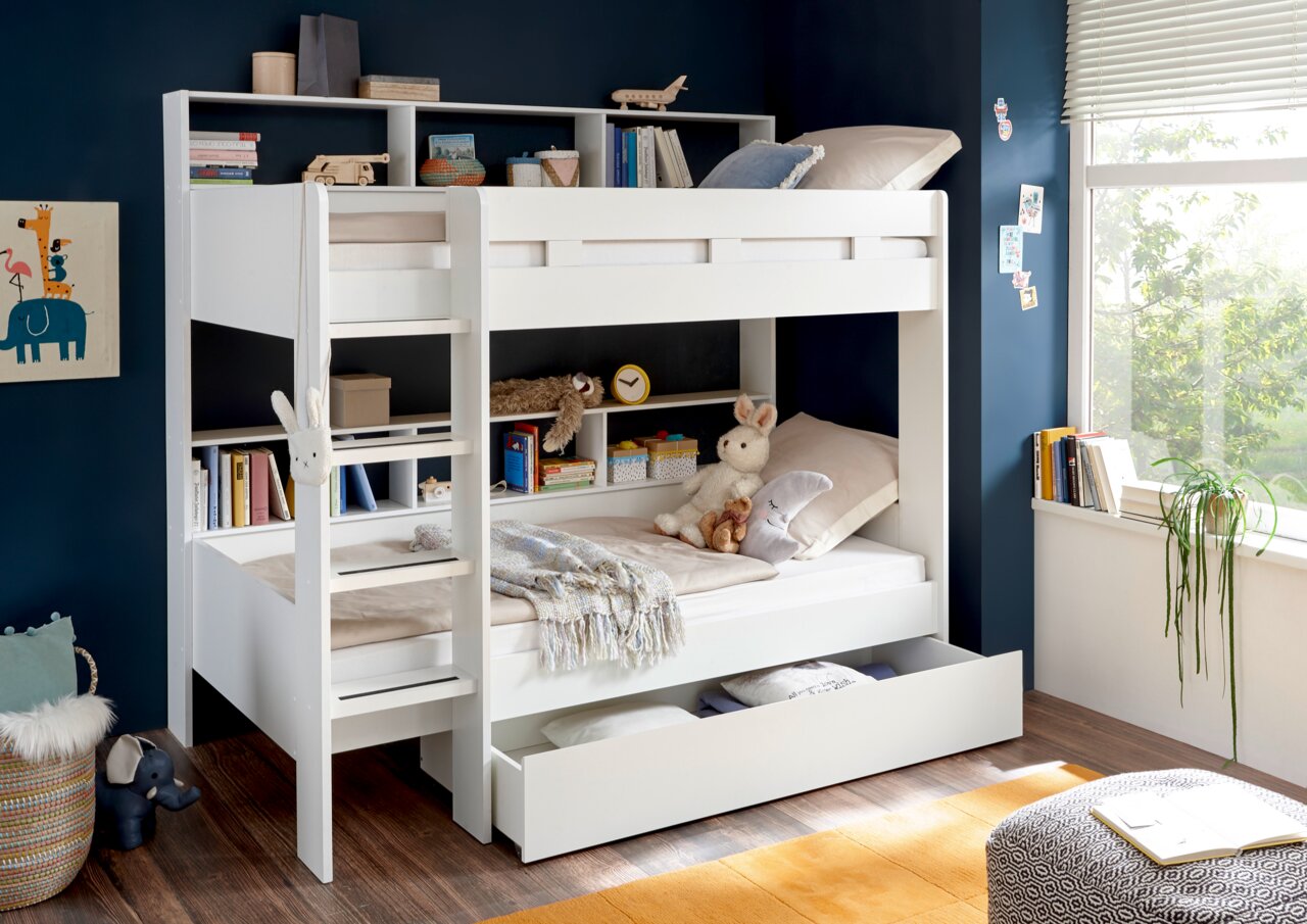 Een wit stapelbed met ingebouwde planken, versierd met speelgoed en boeken, in een lichte kinderkamer met uitzicht op het raam.
