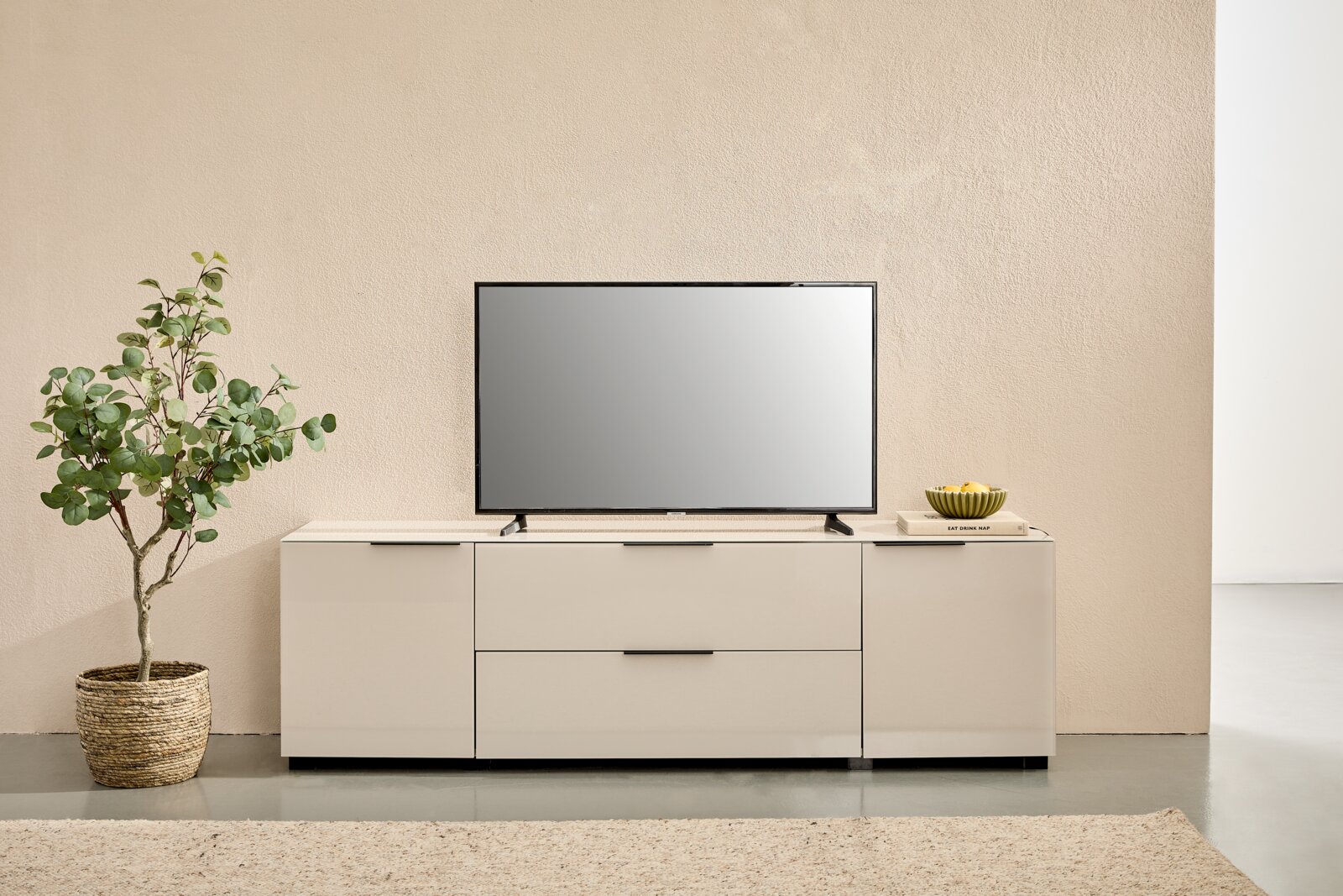 Novel TV-Lowboard MALIN 2.0 Modernes weißes TV-Lowboard mit zwei Schubladen und seitlichen Türen, darauf ein Flachbildfernseher; Frontansicht in minimalistischer Wohnraum-Szene mit Pflanze links und Deko rechts