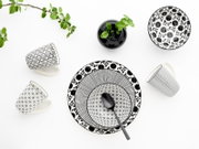 Schwarz-weiß gemustertes Geschirr-Set mit zwei Tassen, Teller und Schale sowie schwarzem Löffel und kleiner Vase mit grünen Blättern, Draufsicht von oben auf weißem Hintergrund