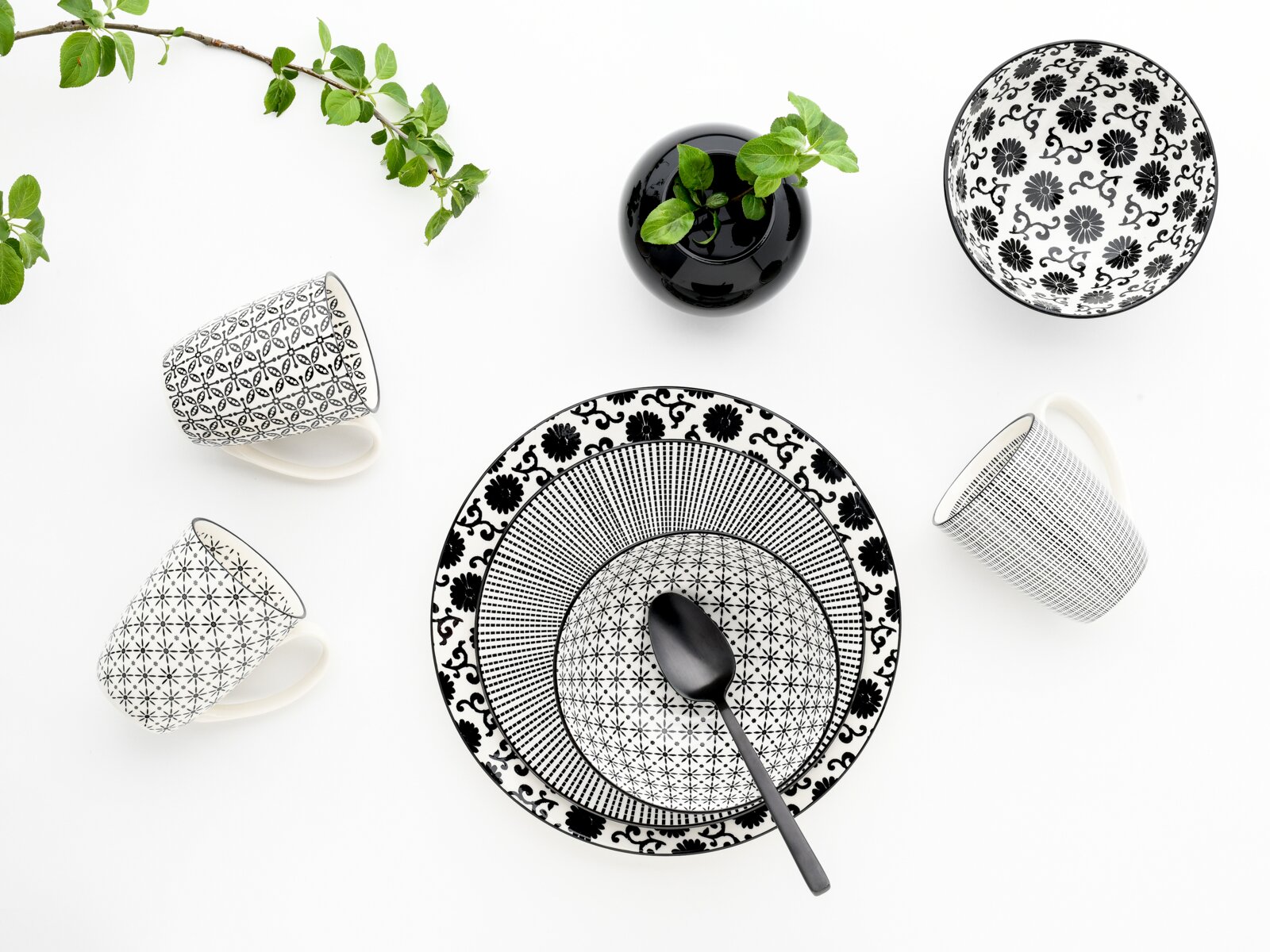 Schwarz-weiß gemustertes Geschirr-Set mit zwei Tassen, Teller und Schale sowie schwarzem Löffel und kleiner Vase mit grünen Blättern, Draufsicht von oben auf weißem Hintergrund