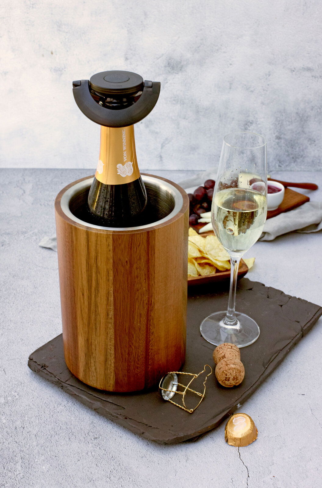 Holz-Weinkühler mit Sektflasche, seitliche Perspektive, daneben ein gefülltes Sektglas und Snacks.