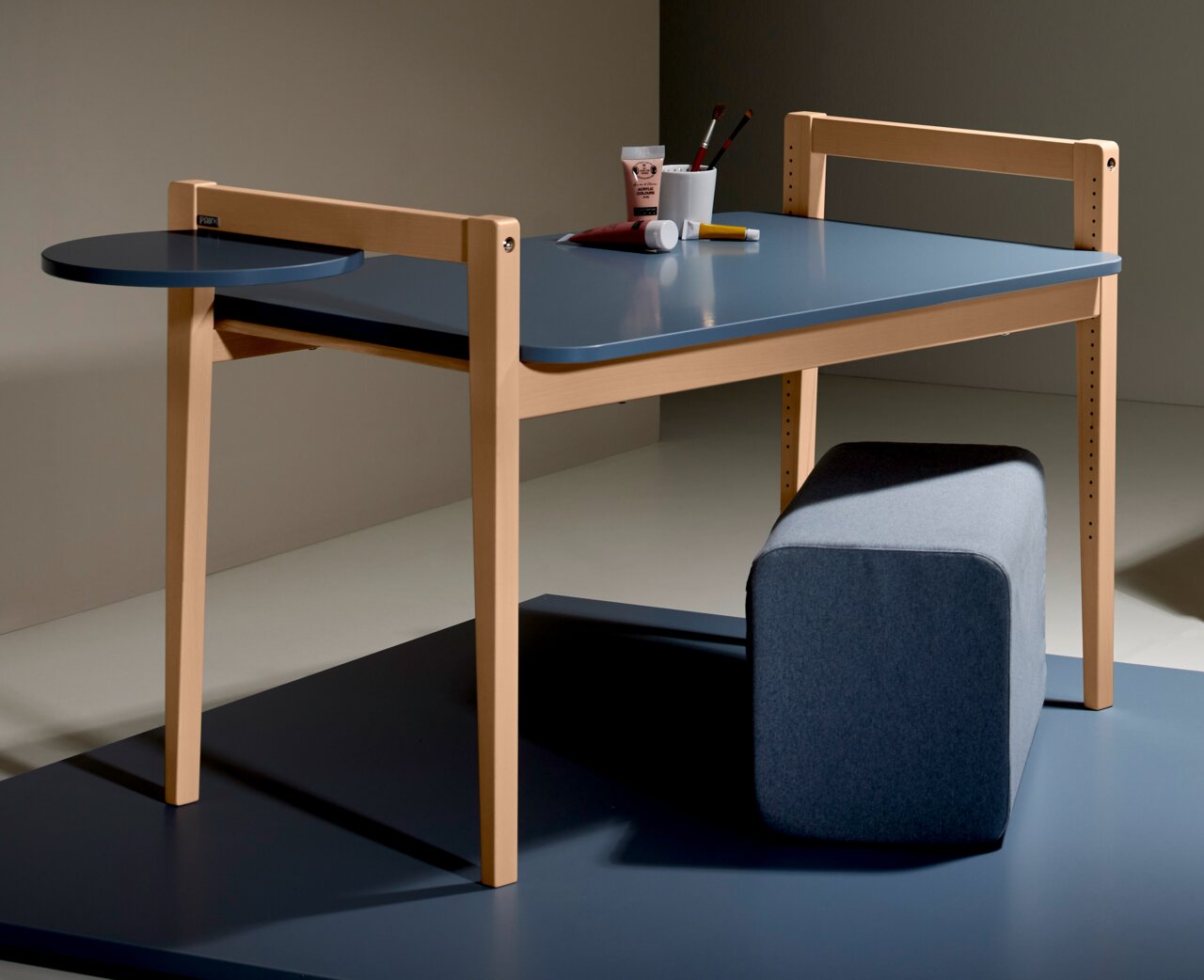 Tischplatte Tiny Table in seitlicher Perspektive mit blauer Oberfläche und hellem Holzrahmen, ergänzt durch eine runde Ablagefläche und einen grauen Hocker.