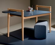Het tafelblad van de Tiny Table in zijaanzicht, met een blauw oppervlak en een licht houten frame, aangevuld met een rond opbergvlak en een grijze kruk.