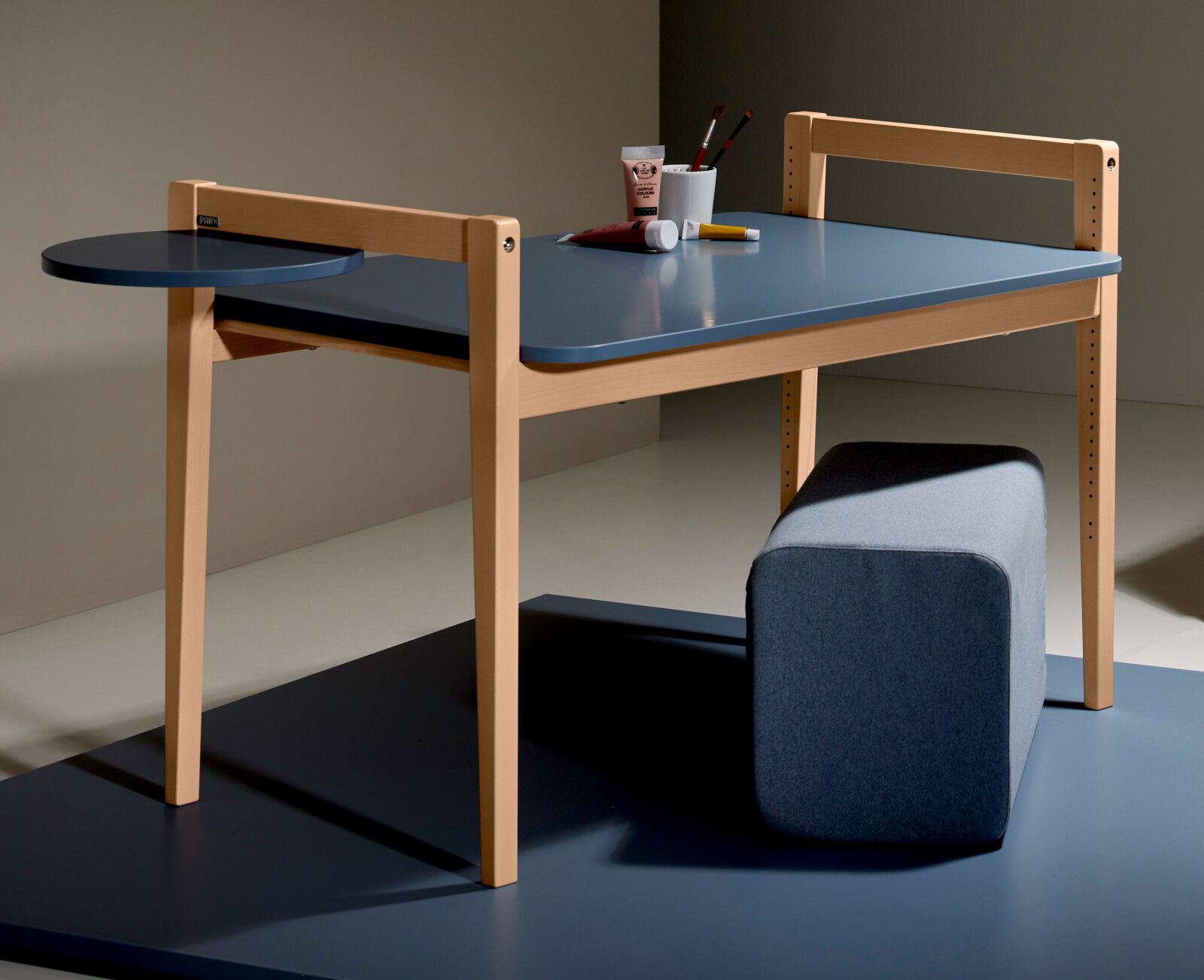Het tafelblad van de Tiny Table in zijaanzicht, met een blauw oppervlak en een licht houten frame, aangevuld met een rond opbergvlak en een grijze kruk.
