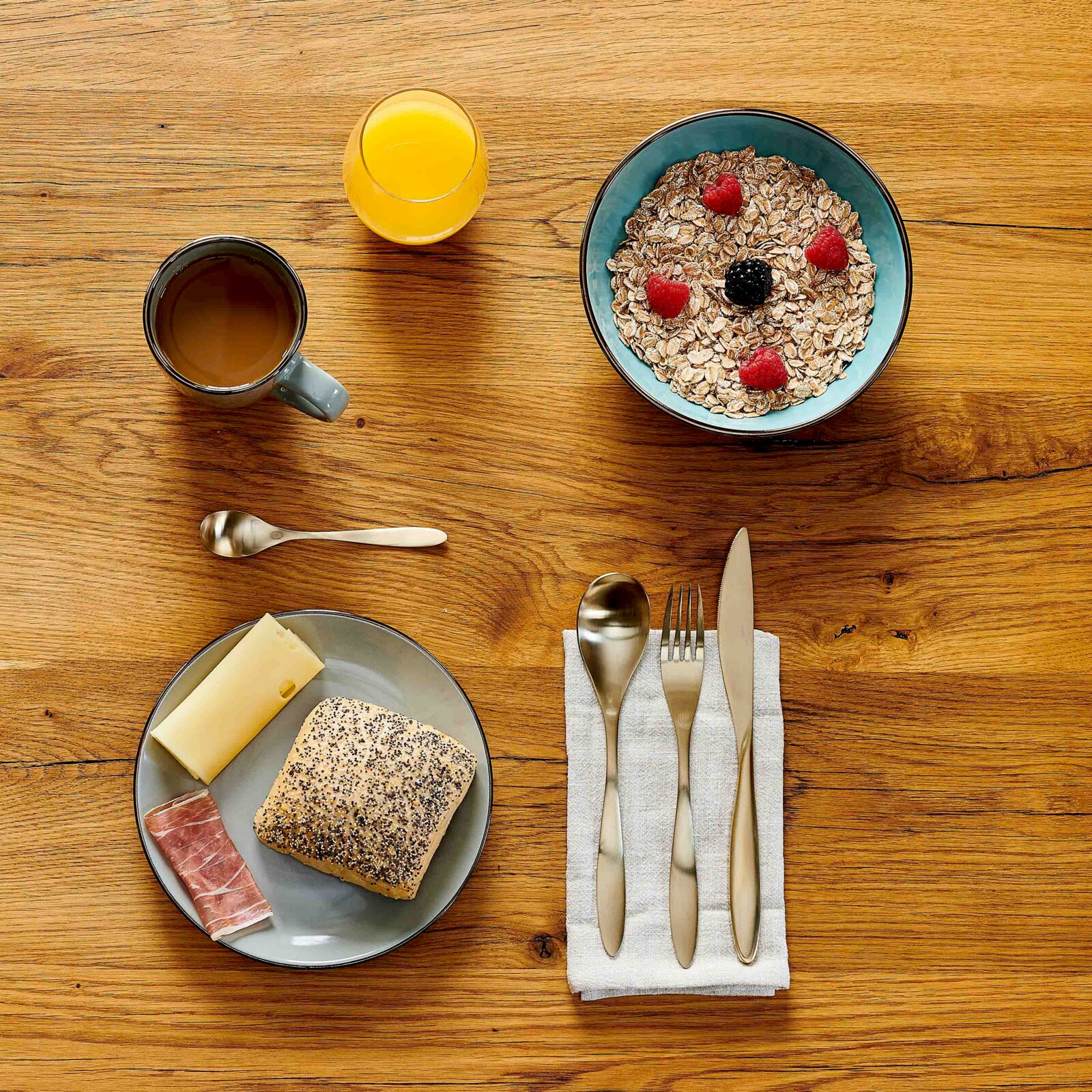 Draufsicht auf einen Frühstückstisch aus Holz mit Schüssel Müsli und Beeren, Tasse Kaffee, Glas Orangensaft sowie Teller mit Brot, Käse und Schinken und Besteck auf Serviette