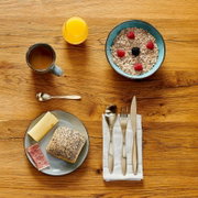 Draufsicht auf einen Frühstückstisch aus Holz mit Schüssel Müsli und Beeren, Tasse Kaffee, Glas Orangensaft sowie Teller mit Brot, Käse und Schinken und Besteck auf Serviette