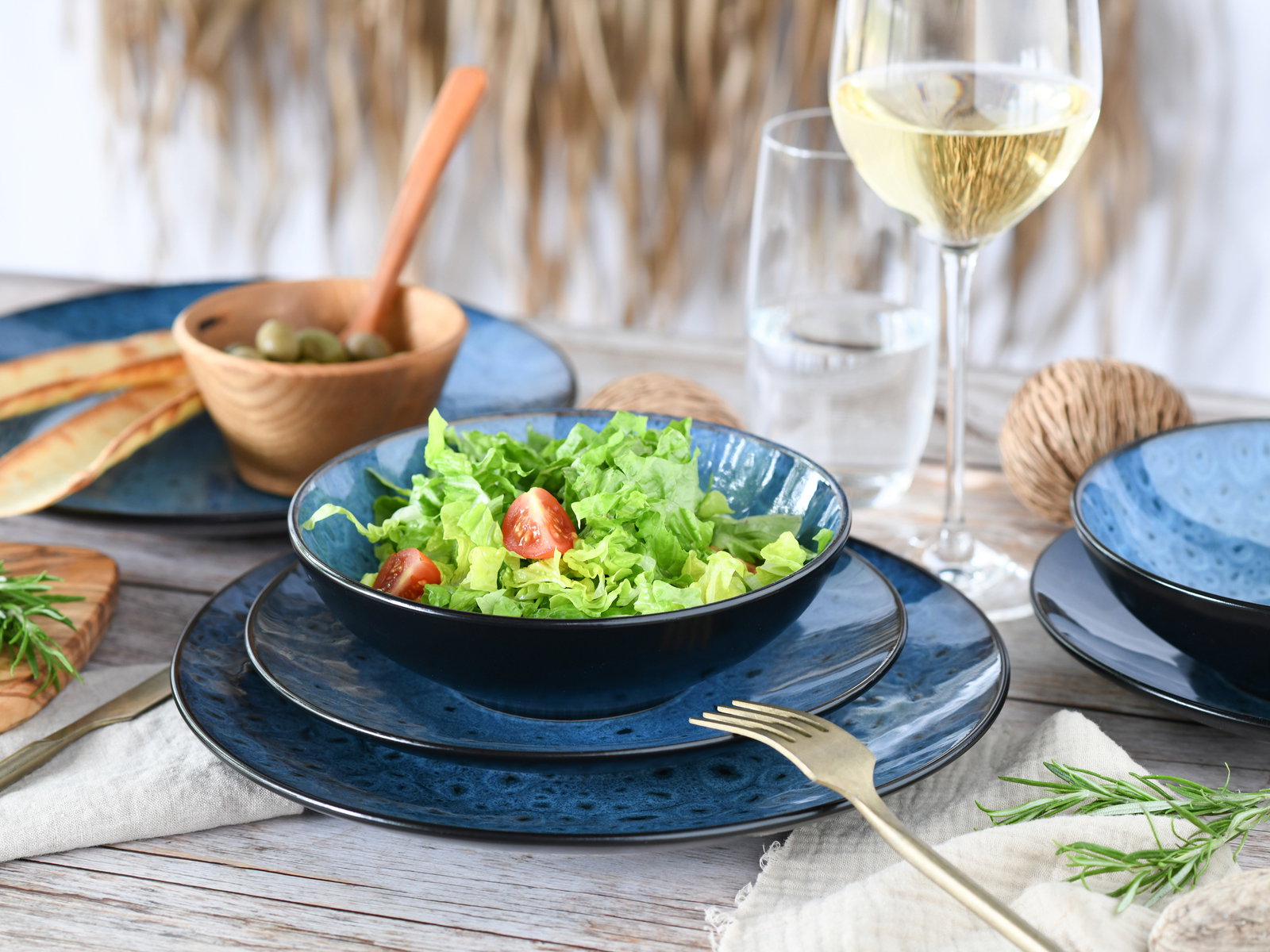 Elegantes Tellerset in Blau mit Salat und Tomaten, von oben betrachtet, auf einem gedeckten Tisch mit Weinglas und Holzschale.