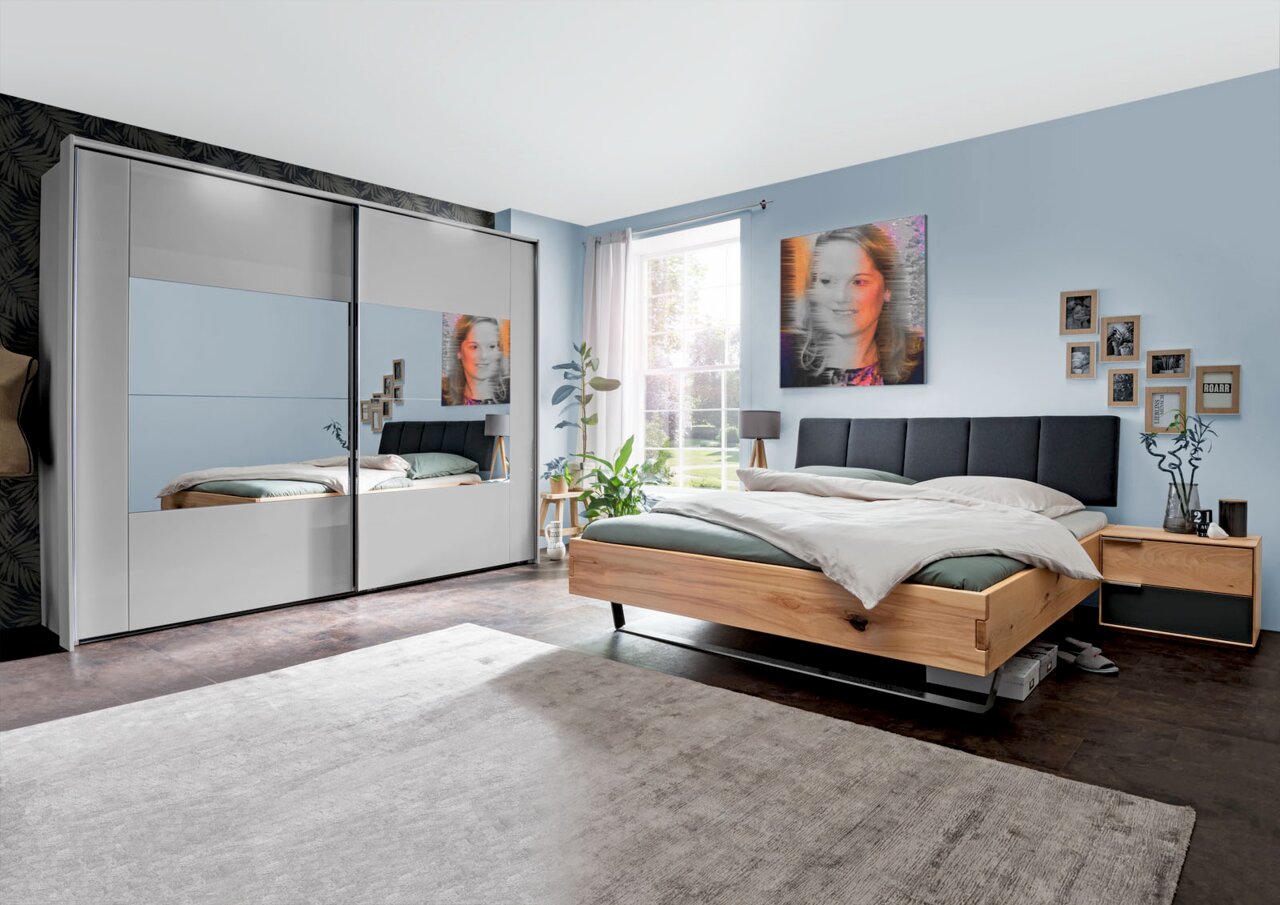 Schlafzimmer mit modernem Schwebetürenschrank in Weiß mit Spiegeln, daneben ein Bett mit Holzrahmen und grauem Kopfteil, seitliche Perspektive.