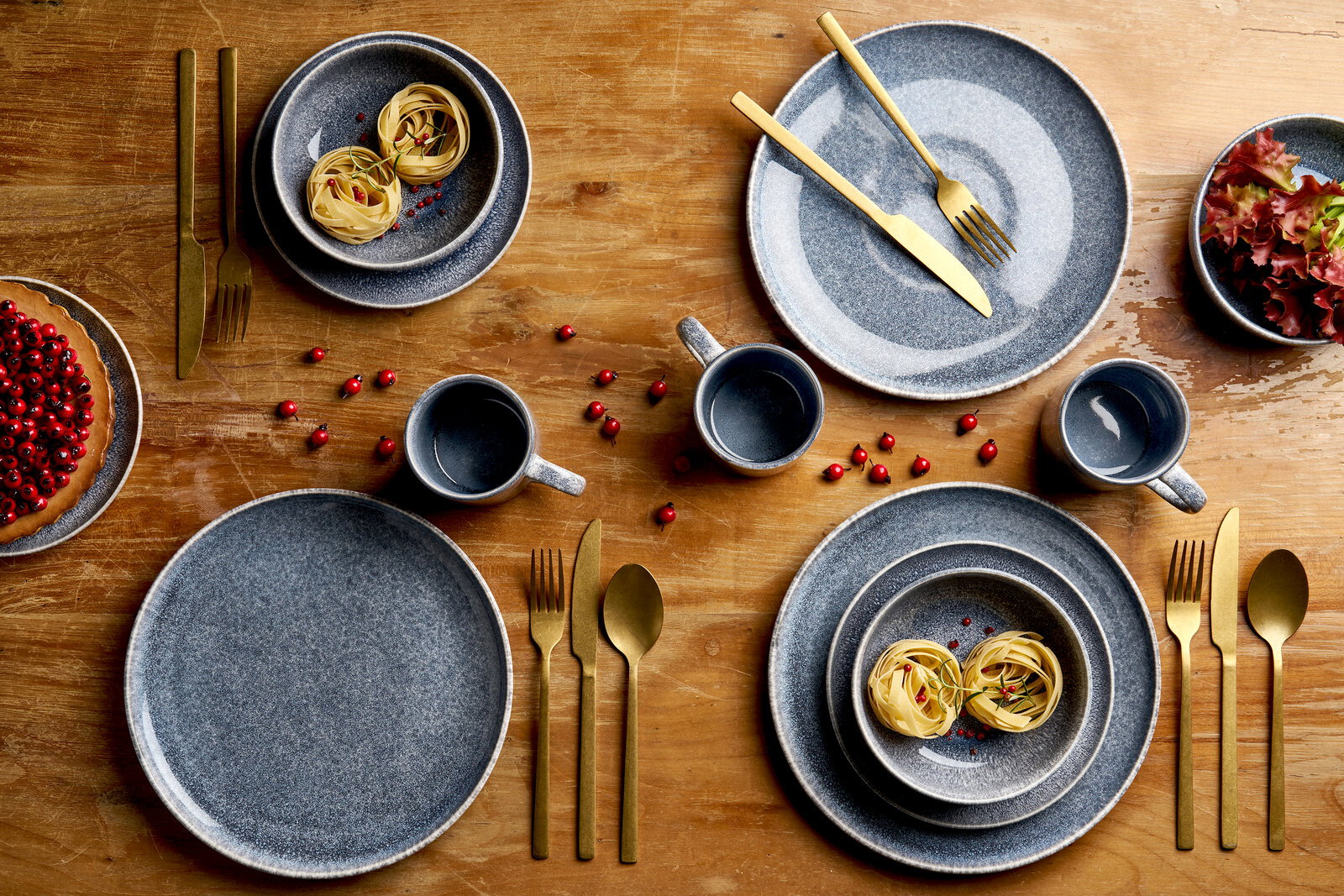 Draufsicht auf ein stilvolles Tischgedeck mit grauem, gesprenkeltem Keramikgeschirr (Teller, Schalen, Tassen) und goldfarbenem Besteck auf Holztisch, dekoriert mit roten Beeren, Pasta-Nestern, Salat und Kuchen
