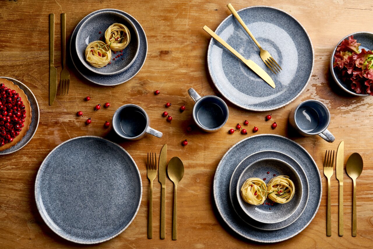 Draufsicht auf ein stilvolles Tischgedeck mit grauem, gesprenkeltem Keramikgeschirr (Teller, Schalen, Tassen) und goldfarbenem Besteck auf Holztisch, dekoriert mit roten Beeren, Pasta-Nestern, Salat und Kuchen
