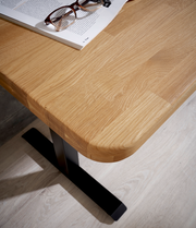 Nahaufnahme eines E-Schreibtischs mit einer Holzoberfläche und einem schwarzen Metallfuß, von oben betrachtet. Auf dem Tisch liegt eine Brille auf einem geöffneten Buch.