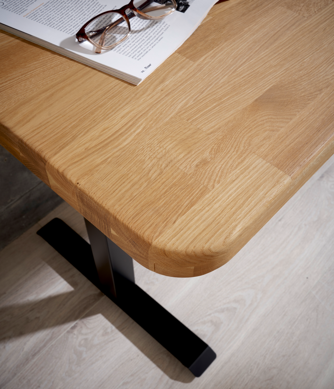 Nahaufnahme eines E-Schreibtischs mit einer Holzoberfläche und einem schwarzen Metallfuß, von oben betrachtet. Auf dem Tisch liegt eine Brille auf einem geöffneten Buch.