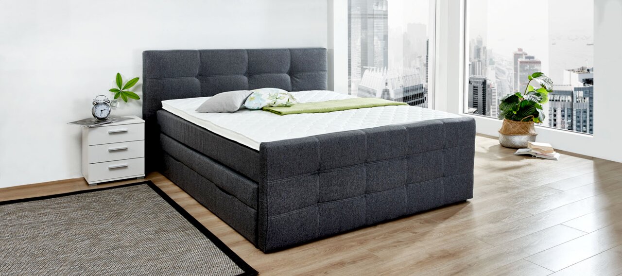 Graues Boxspringbett in modernem Schlafzimmer mit Stadtblick, seitliche Perspektive